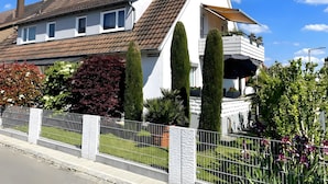 A two-story house with a sloped roof, a balcony with potted plants, and a white fence.