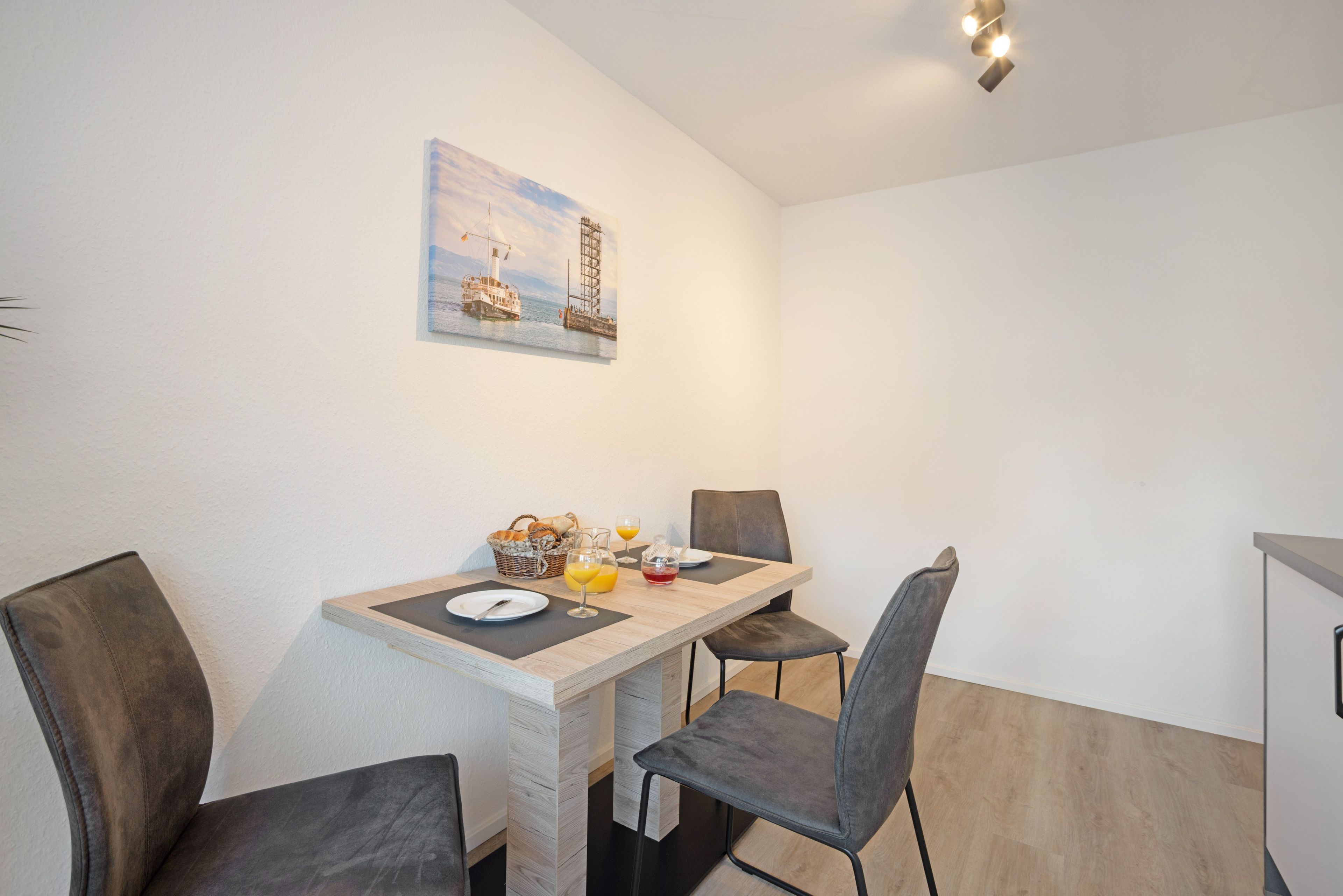 En matplats med ett bord dukat för två personer, stolar, en tavla på väggen och en korg med bröd.