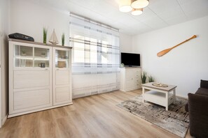 Una sala moderna con suelo de madera, un mueble blanco con puertas de cristal, un televisor de pantalla plana, una pala de madera en la pared y una pequeña mesa con una planta.
