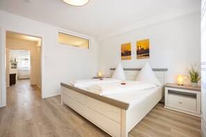 A modern bedroom with a wooden bed, bedside tables, and a nightstand. There are two paintings on the wall, a window with blinds, and a door leading to another room.