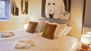 A neatly made bed with pillows and towels, a large painting on the wall, and a bedside table with a lamp.