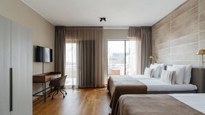 Une chambre d’hôtel avec un plancher en bois, un bureau avec une chaise, deux lits, une télévision et de grandes fenêtres avec des rideaux.