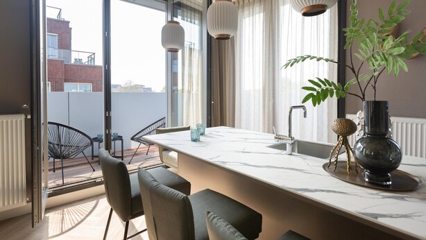 Une cuisine moderne dotée d’un grand îlot central, d’un coin repas avec des chaises et offrant une vue sur la ville grâce à de grandes fenêtres.