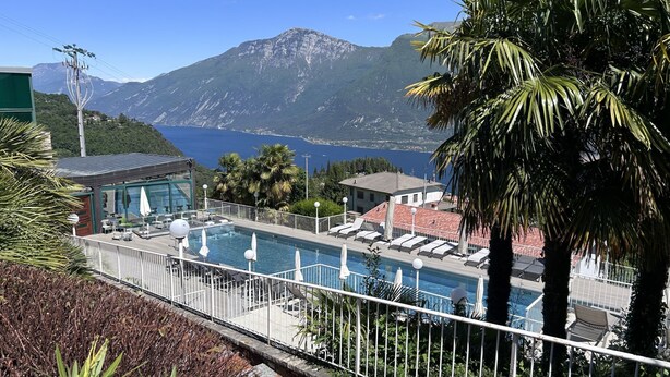 Ein Poolbereich mit Liegestühlen, ein Gebäude mit rotem Dach und eine Bergkulisse.