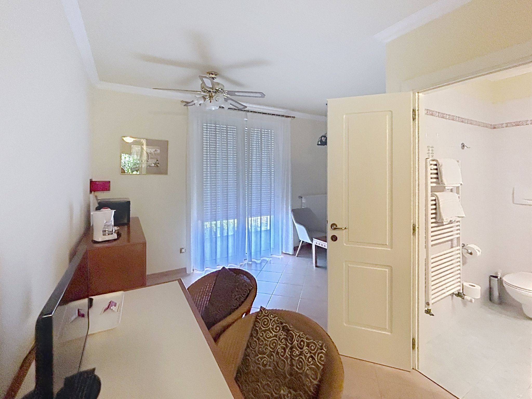 A hotel room with a dining area, a ceiling fan, a bathroom with a toilet and towel rack, and a door leading to another room.