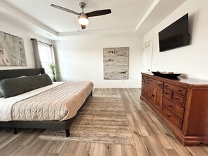 A bedroom with a bed, a wooden dresser, a ceiling fan, a painting on the wall, and a television mounted on the wall.