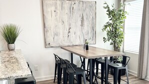 A modern kitchen with a granite countertop, black bar stools, a wooden dining table, and a large abstract painting on the wall.