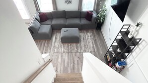 A staircase leading to a living area with a grey sofa, a small ottoman, a wooden floor, and a TV mounted on the wall.