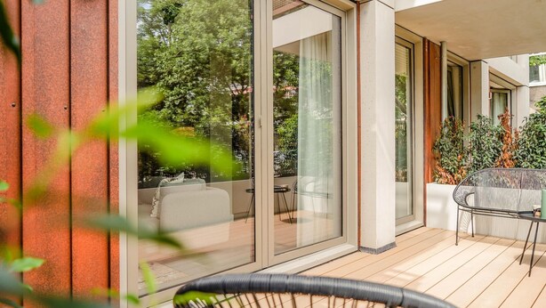 A modern outdoor patio with a wooden deck, a glass door leading to a room, and a wicker chair. - Sustainable Stay with Terrace (Den Haag)