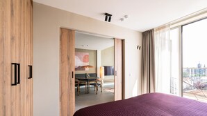 A modern hotel room with a large bed, a wooden wardrobe, a dining area with a table and chairs, and a large window offering a city view.