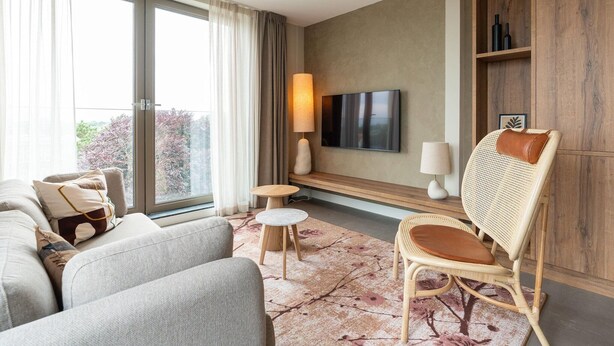 A modern living room with a sofa, armchair, and a small round table. A flat-screen TV is mounted on the wall, and there is a wooden bookshelf to the right.