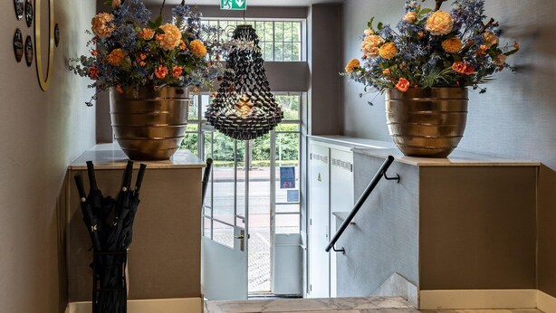 Een hotel lobby met twee grote bloemstukken op voetstukken, een hanglamp en uitzicht op buiten door een glazen deur.