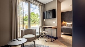 A modern hotel room with a large window, a desk, a comfortable chair, a bed, and a flat-screen TV. - Modern Stay in Historic Delft (Den Haag)