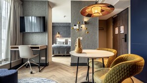 A modern hotel room with a flat-screen TV, a desk, a comfortable bed, a dining area with a round table and cushioned chairs, and a large window with curtains. - Modern Stay in Historic Delft (Den Haag)