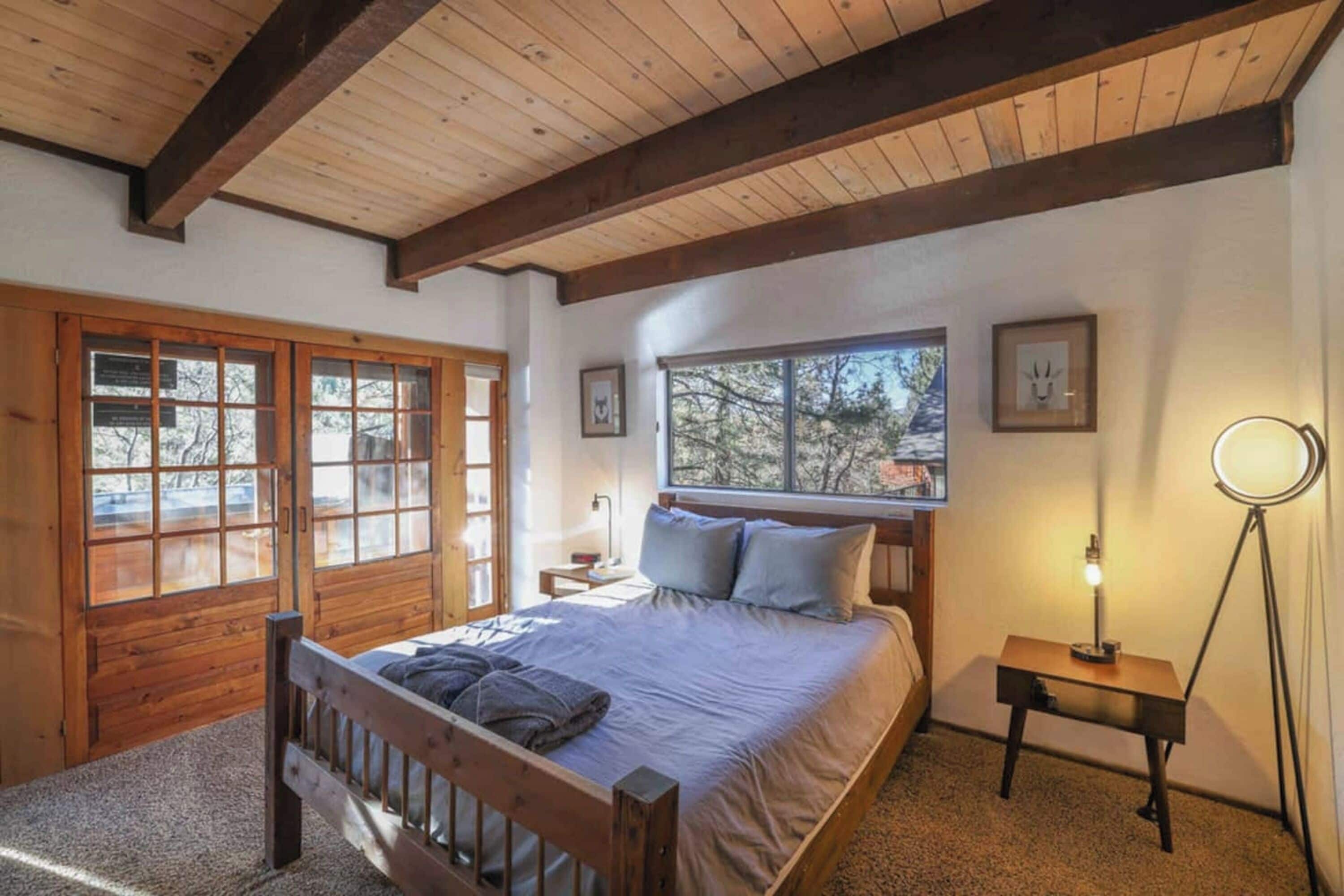 Un dormitorio con cama de madera, una mesita de noche, una lámpara de pie y ventanas amplias.