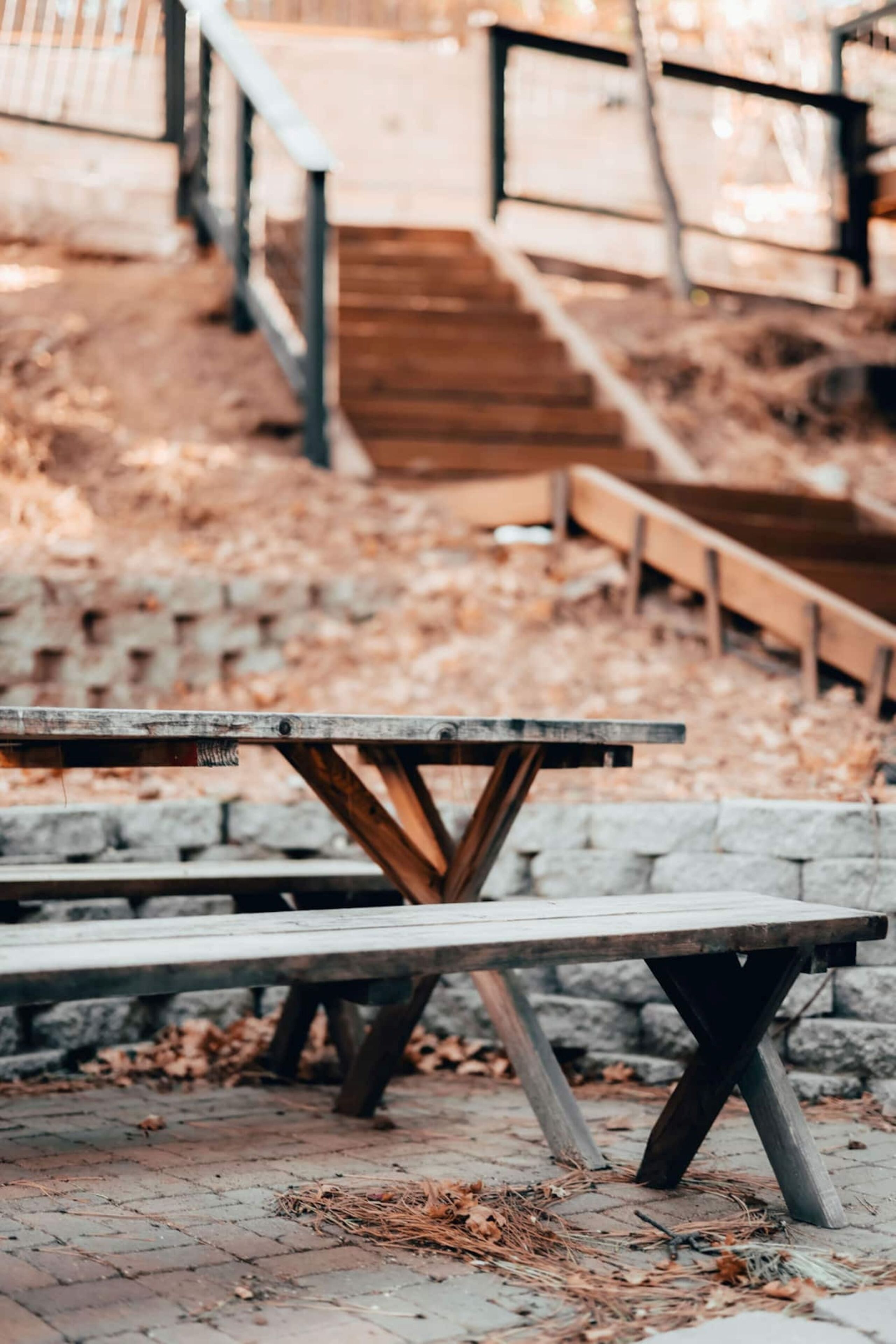 Une table de pique-nique en bois, dotée d’un cadre en métal, est placée sur une zone pavée de briques, parsemée de feuilles.