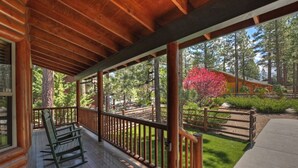 A wooden porch with rocking chairs, a wooden fence, and a view of a forested area with a red flowering tree. - Teakwood Cozy Wood Cabin with Fireplace (Big Bear Lake)