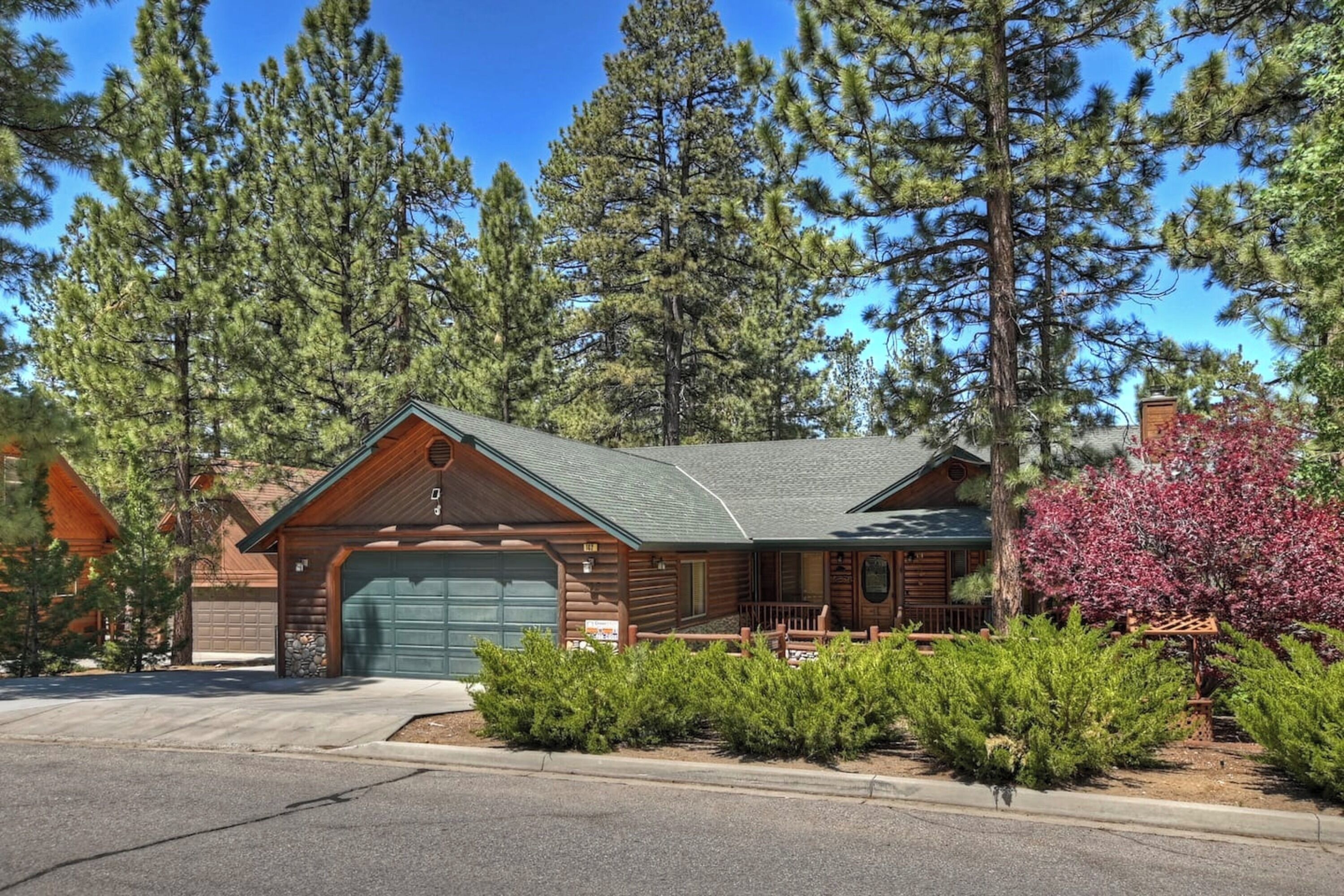 A log cabin with a green garage, surrounded by pine trees and a small garden.