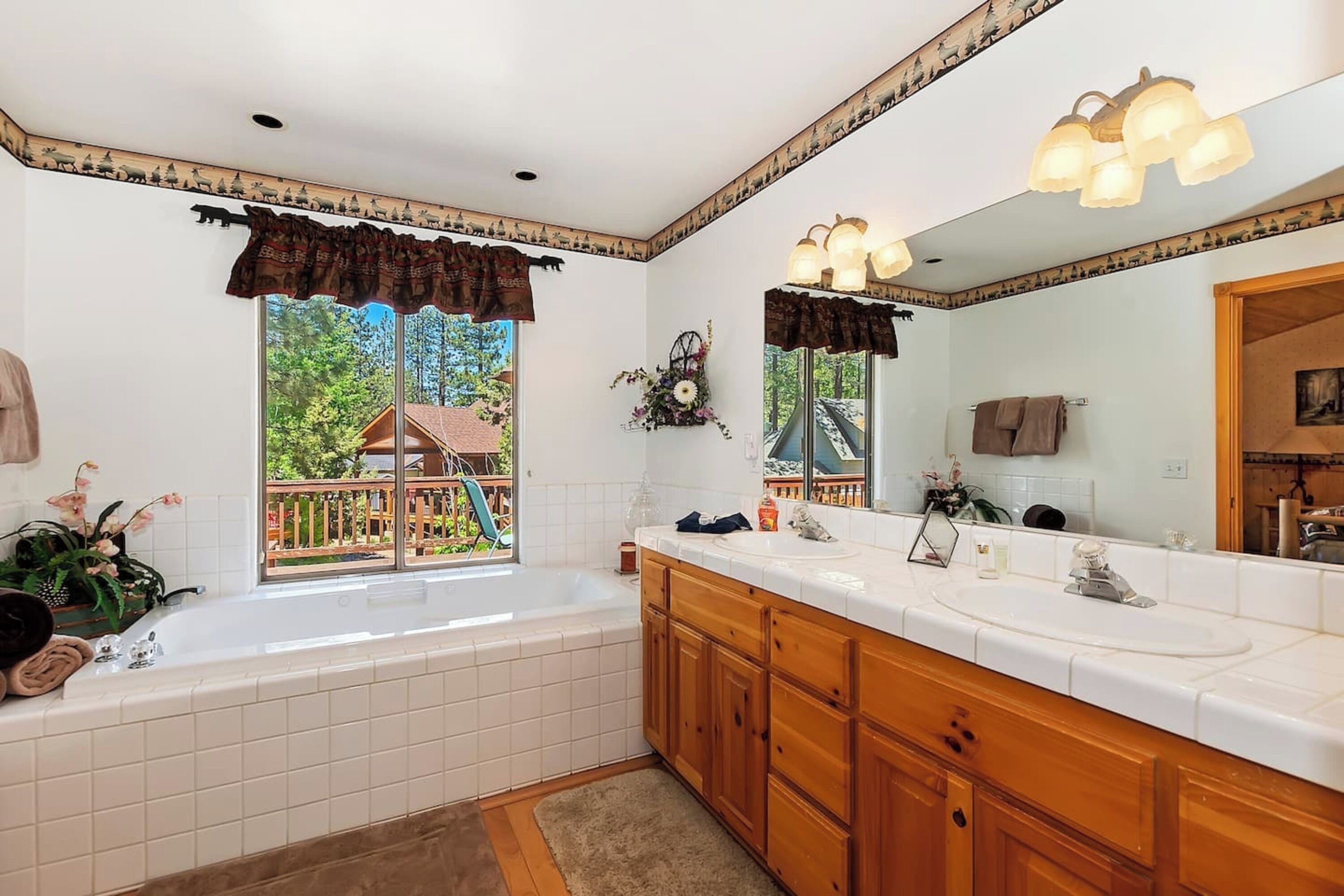 A bathroom with a large window, a bathtub, a double sink, wooden cabinets, and a mirror.