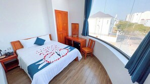 A hotel room with a bed, a desk, a chair, and a large window with a view of buildings outside. - Blue Ocean Hotel Phan Thiet - BAY LUXURY (Phu Thuy)
