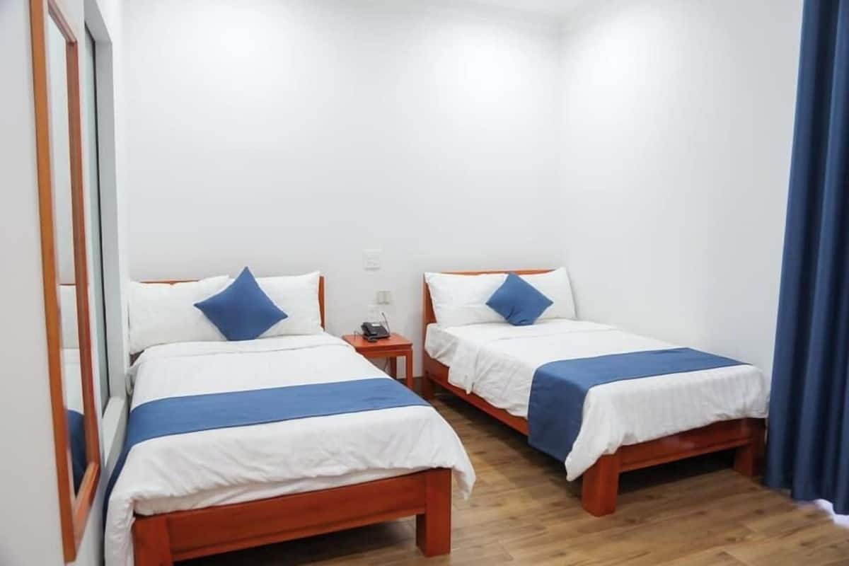 A room with two wooden beds, each with white bedding and blue accents, a small bedside table, and a mirror on the left wall.
