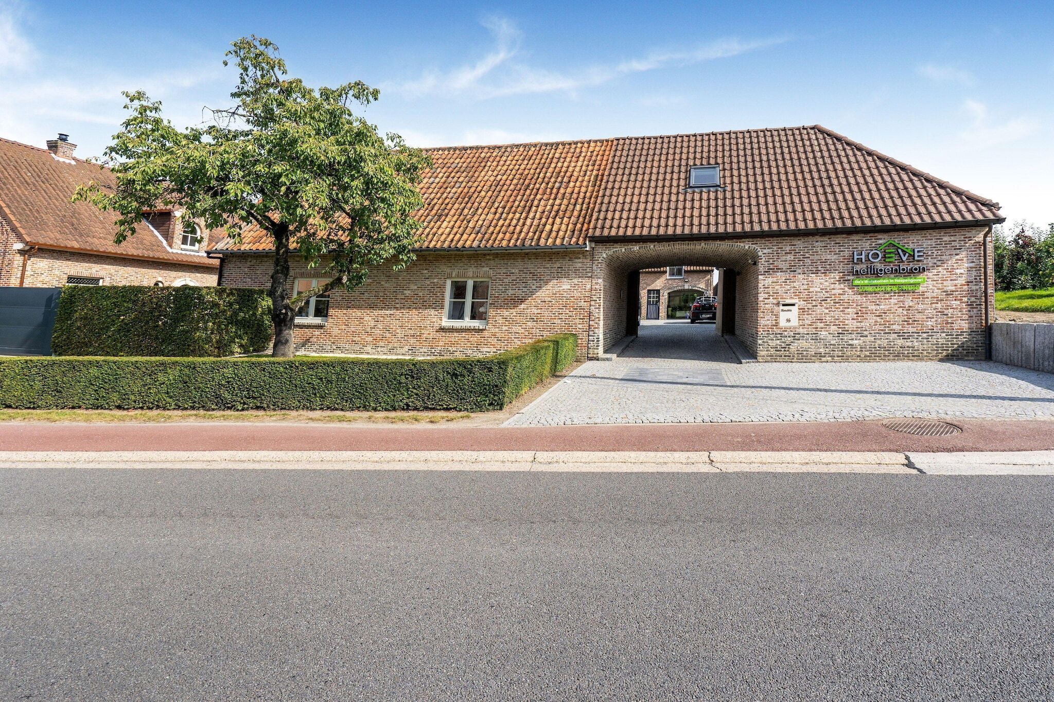 A brick building with a tiled roof, a sign reading "HOVE helligenbron," and a paved entrance.