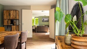 A modern living room with a wooden dining table, brown leather chairs, a grey sofa, and a large window with a view of greenery. - Group home in Andijk with garden room (Andijk)