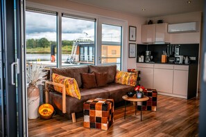 A modern living room with a sofa, ottoman, and a small table. There is a kitchen area with cabinets and appliances. Large windows provide a view of the outdoors.