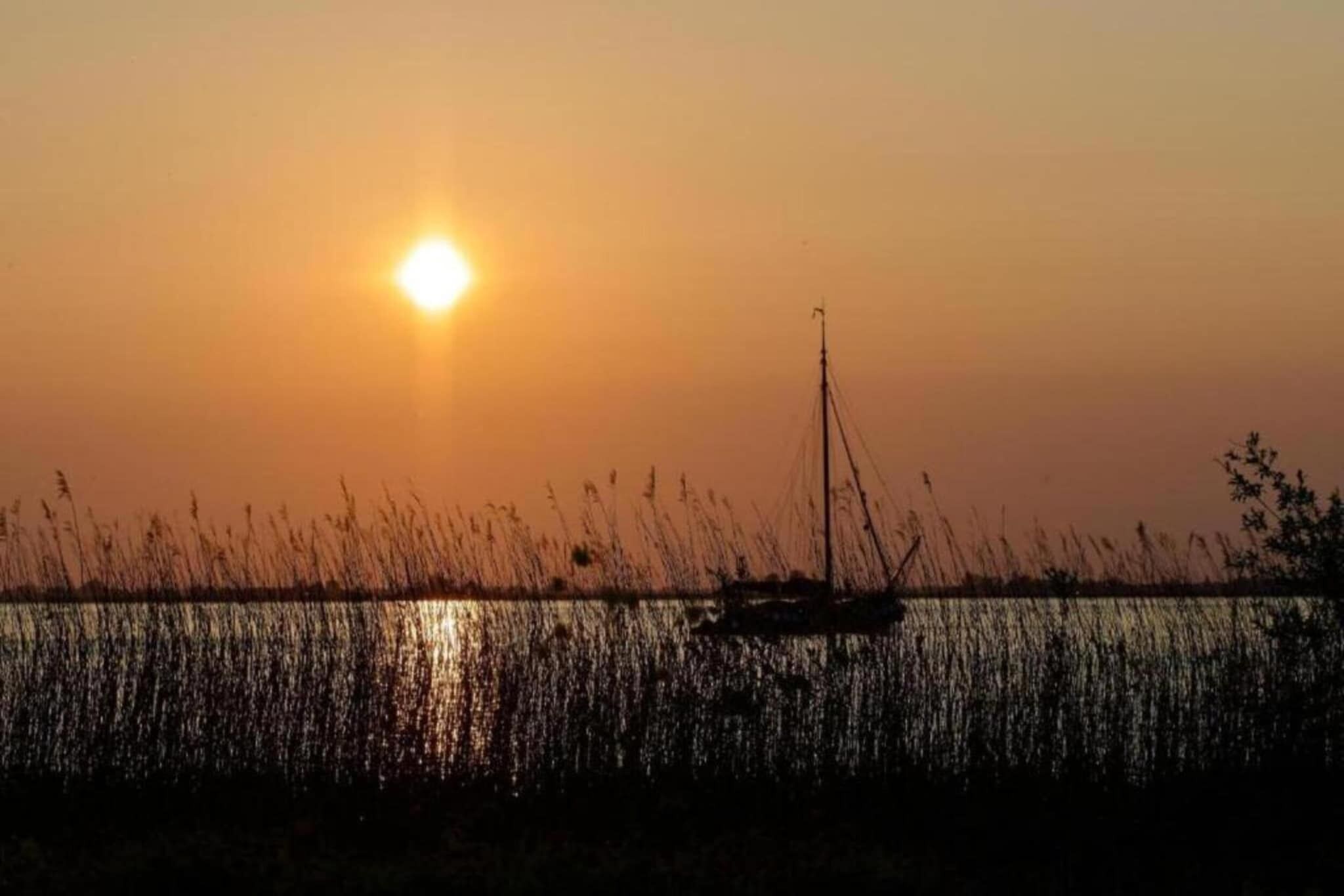 A sunset over a calm body of water with a sailboat and reeds in the foreground.