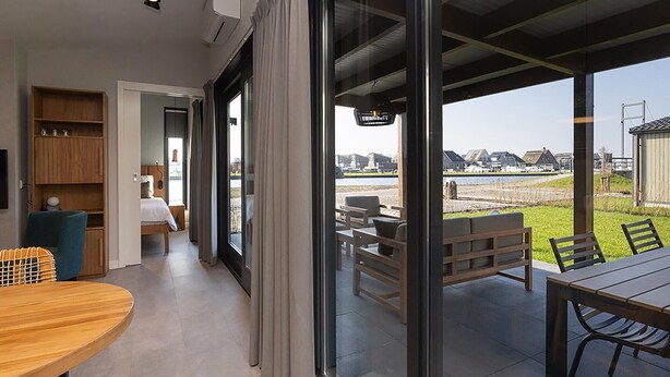 Een moderne hotelkamer met een grote schuifdeur naar een terras. De kamer heeft een houten eettafel, een bank en een inbouwkast.