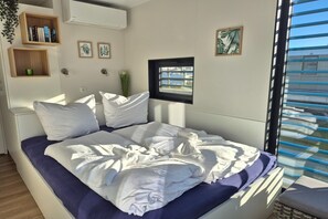 A bed with white and purple bedding, a small window, and wall-mounted shelves with books and decorative items. - Floating Comfort in Peenemünde (Peenemünde)