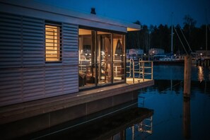 A waterfront houseboat with a glass-enclosed deck, a visible interior with a fireplace, and a dock extending into the water. - Floating Comfort in Peenemünde (Peenemünde)