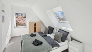 Ein modernes Schlafzimmer mit schrägen Wänden, einem Oberlicht, einem Bett mit vielen Kissen und einem Holzschrank.