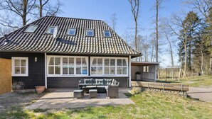 A house with a tiled roof, large windows, a patio with outdoor furniture, and a stone wall.