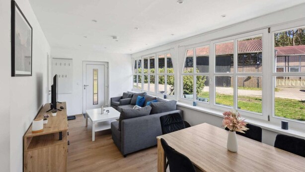 Una sala de estar moderna con un conjunto de sofás, una mesa de comedor y ventanas grandes con vistas al exterior.