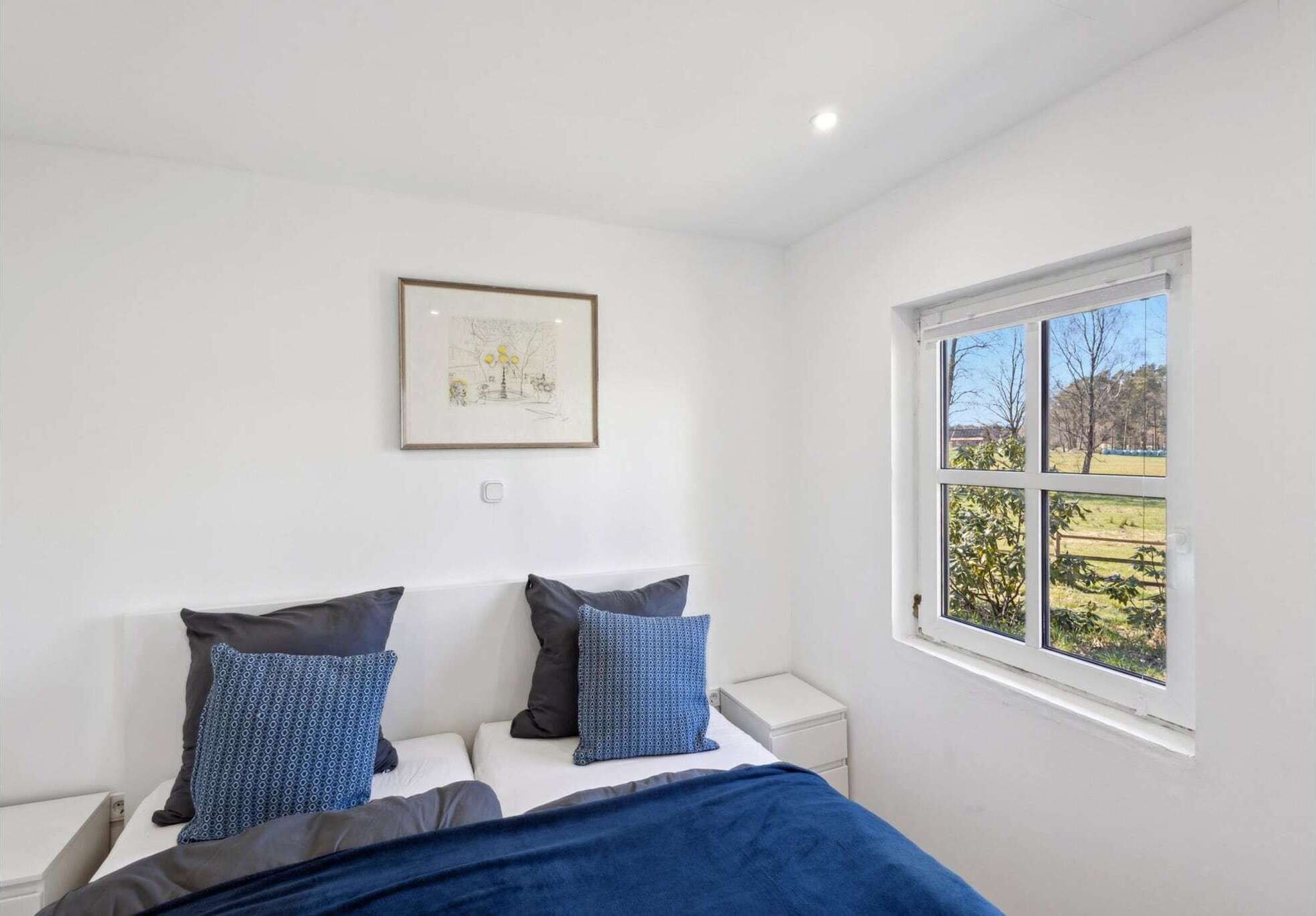 A bedroom with a bed, two pillows, a bedside table, a framed picture on the wall, and a window with a view of greenery.