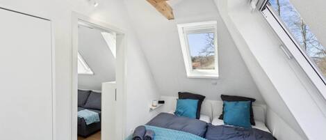 Un dormitorio con techo inclinado, cama con ropa de cama azul y gris, una mesita de noche pequeña y una ventana con vistas al exterior.