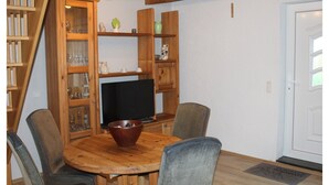 A dining area with a wooden table, chairs, and a television. There are wooden shelves with various items and a bookshelf to the left. - Ferienhaus in Rethwisch (Rethwisch)