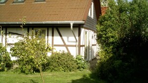 A traditional half-timbered house with a sloped roof, a small porch, and a garden area with various plants and a tree. - Ferienhaus in Rethwisch (Rethwisch)