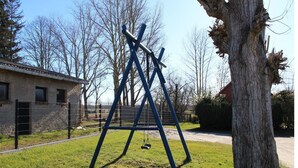 A swing set in a grassy area with a tree and a building in the background. - Ferienhaus in Rethwisch (Rethwisch)