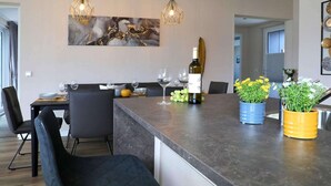 A modern kitchen with a central island, pendant lights, a wine bottle, and potted plants.