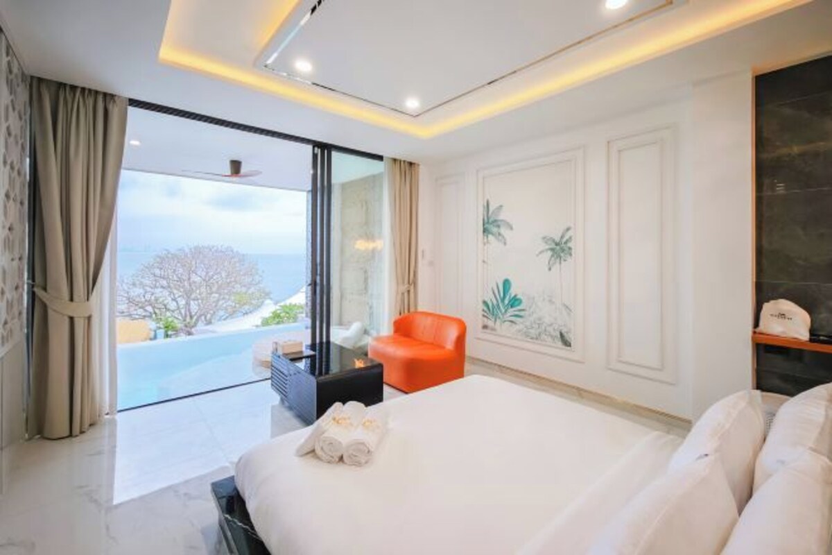 A modern hotel room with a large bed, a seating area with a red chair, a small table, and a view of the outdoors.