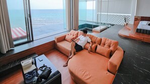 Une chambre d’hôtel moderne avec une grande fenêtre donnant sur la mer, un canapé orange moelleux, une petite table avec un téléphone et un sac, et un espace nuit séparé avec un lit et une petite table.