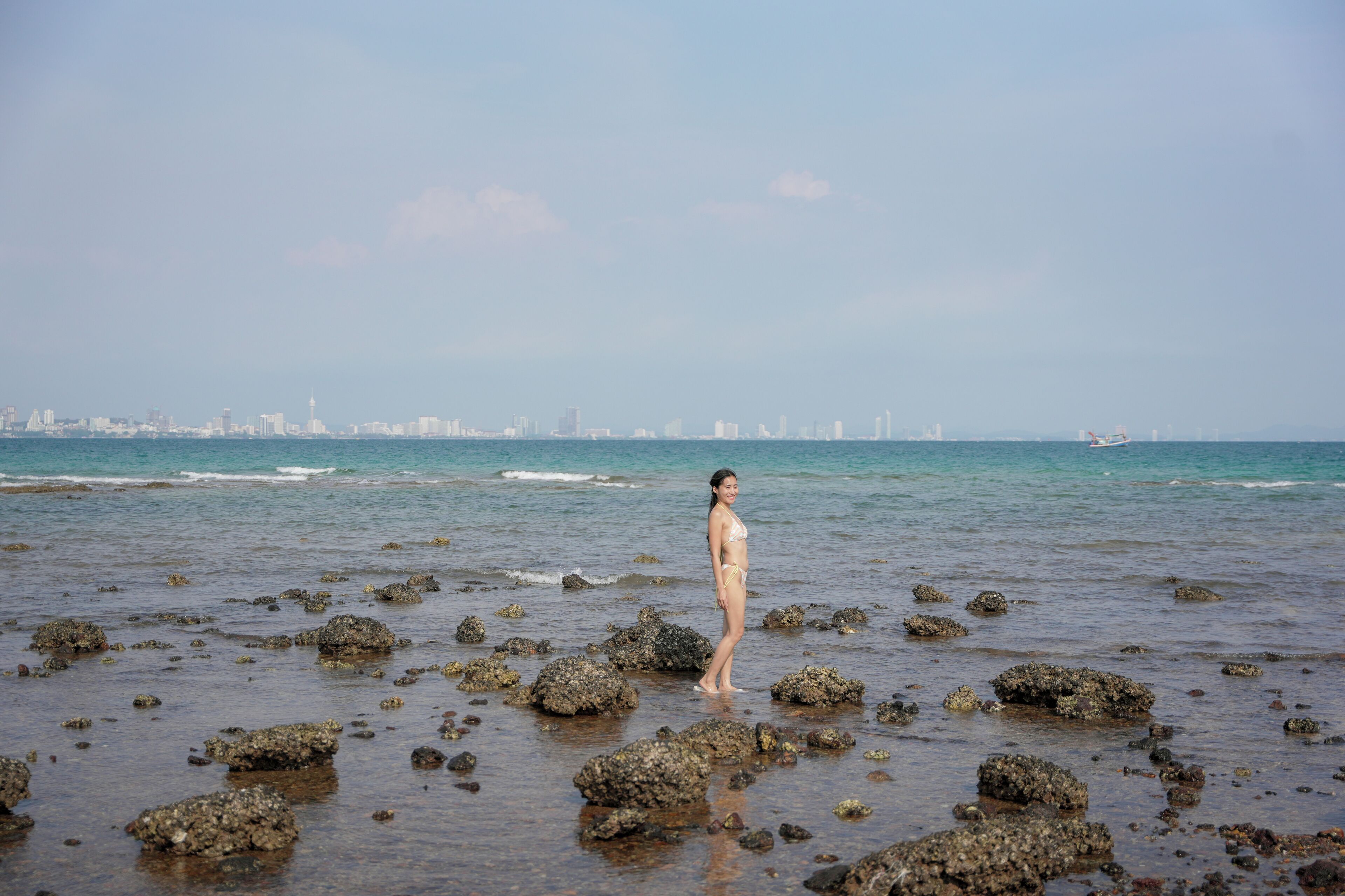 Una persona de pie en una playa rocosa, con un horizonte urbano al fondo.