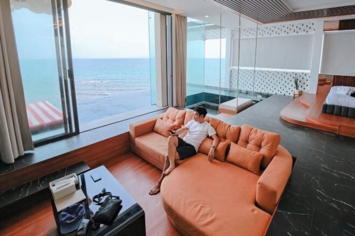 A modern hotel room with a large window offering a view of the ocean, a plush orange sofa, a small table with a bag, and a bed with a white headboard.