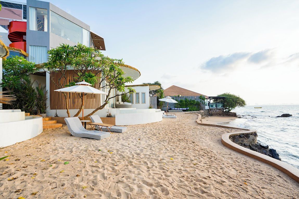 A beachfront property with a sandy beach, lounge chairs, umbrellas, and a modern building with a curved roof.