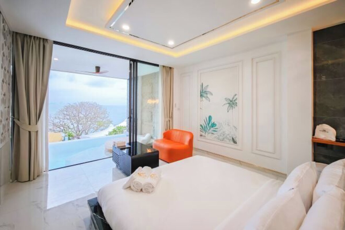 A modern hotel room with a large bed, a seating area with a red chair, a small table, and a view of the outdoors.