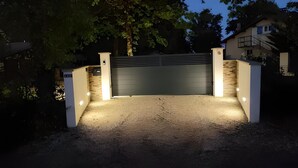 Ein befestigter Auffahrtsweg mit geschlossenem Garagentor, umgeben von einer weißen Mauer mit Steinverzierungen und beleuchtet durch Bodeneinbaulichter.