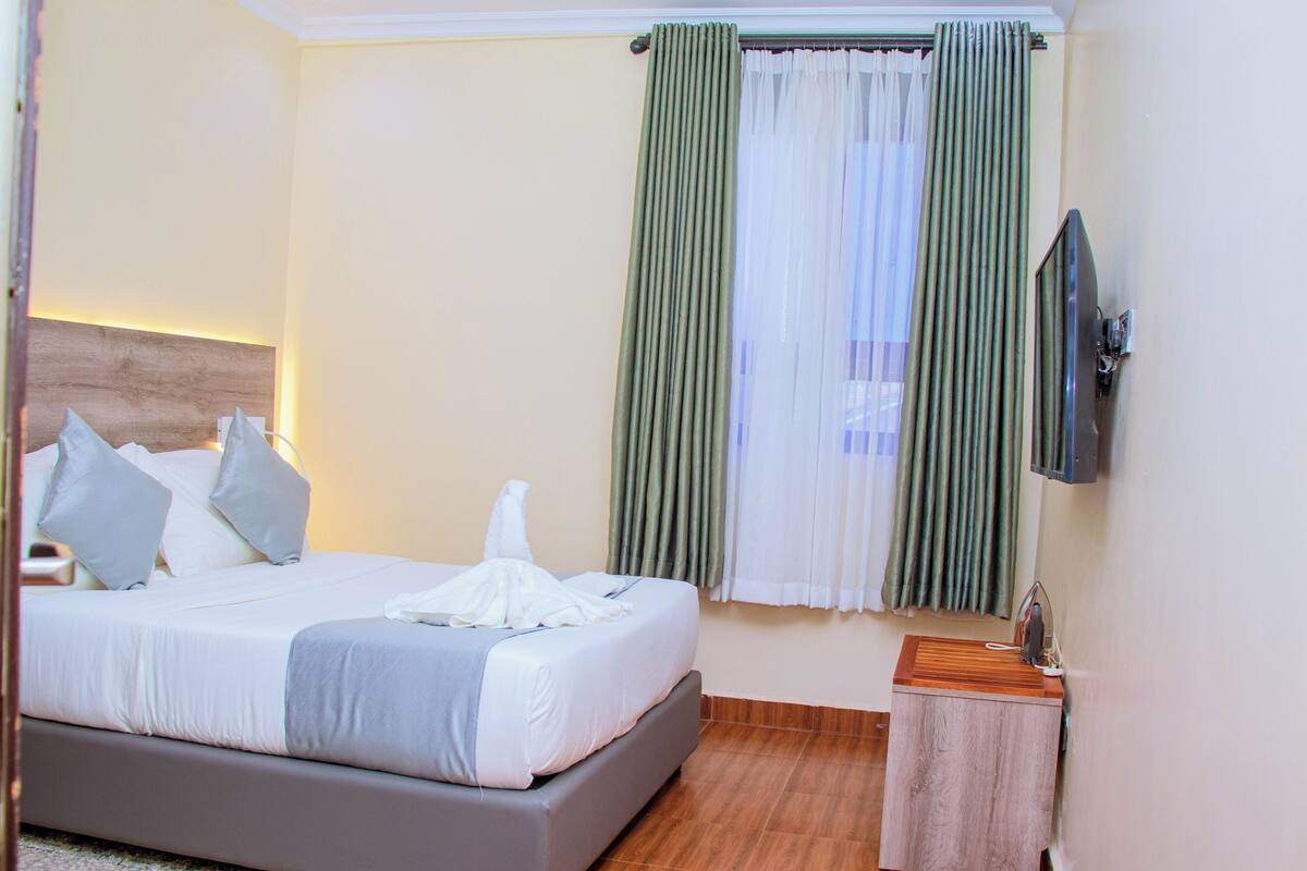 A neatly made bed with pillows, a bedside table, a flat-screen TV, and a window with curtains.