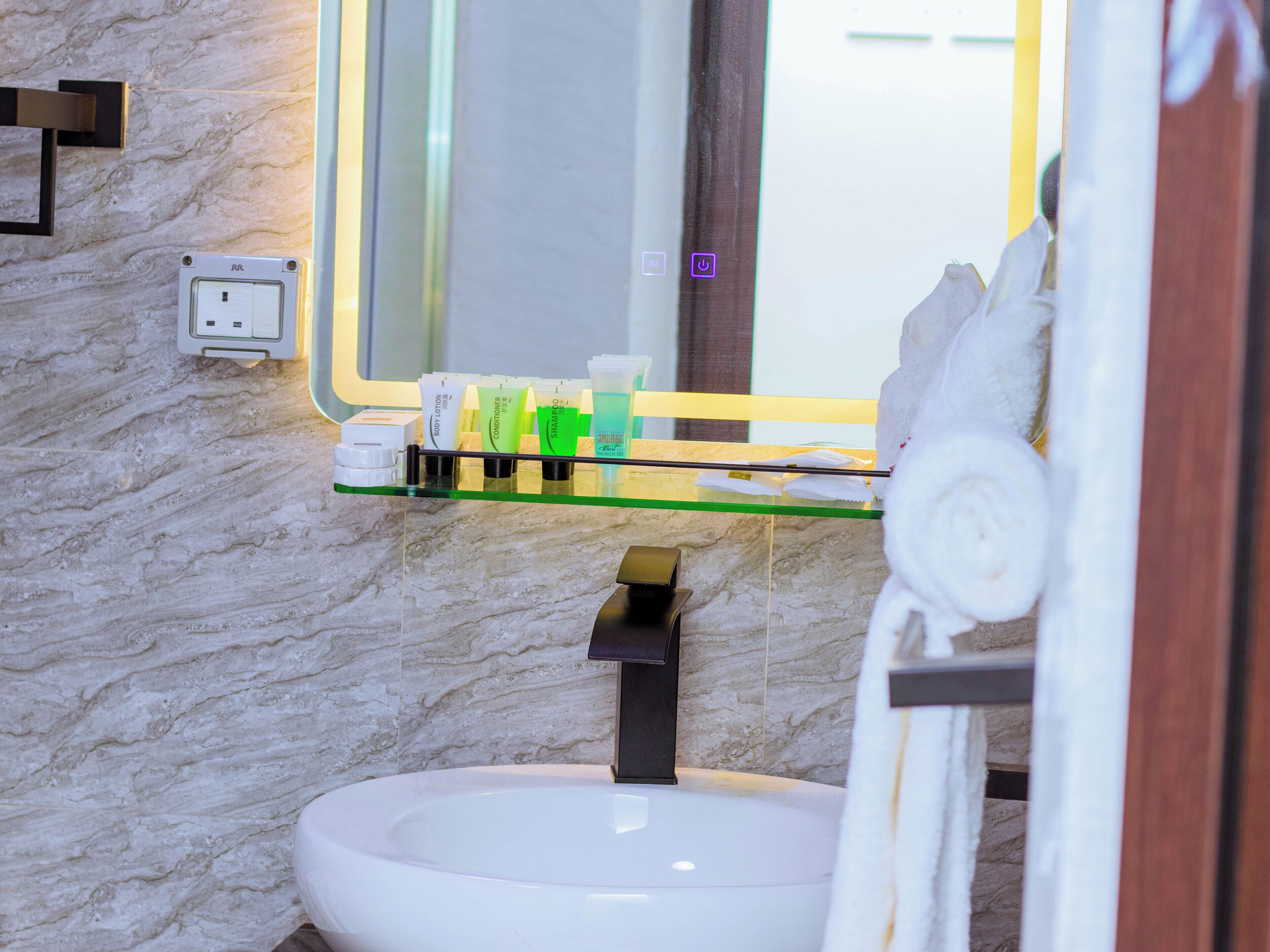 A bathroom with a marble wall, a glass shelf with toiletries, a white sink, and a mirror.
